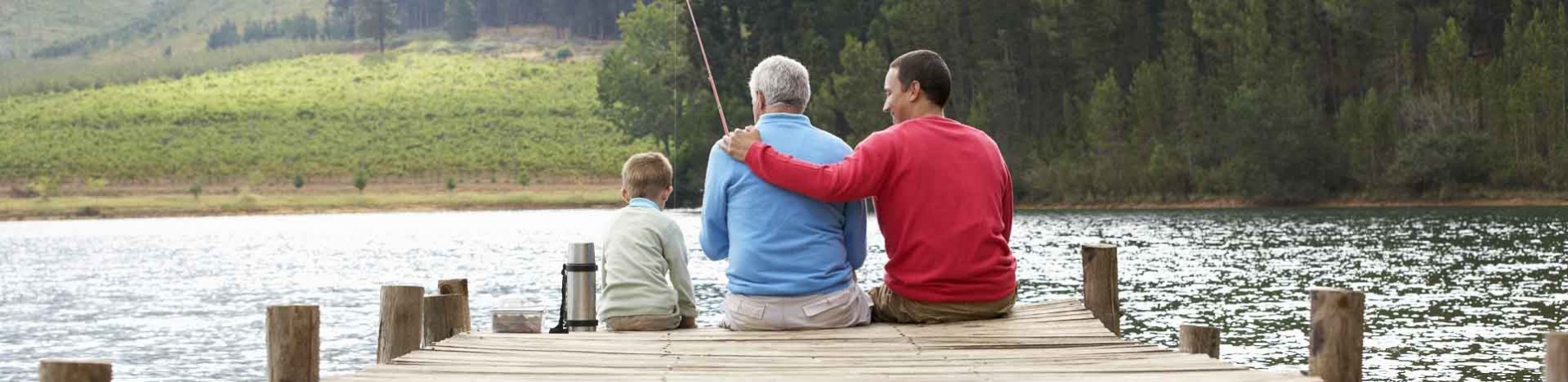 Three male generations sitting at the end of a dock at a beautiful lake while fishing.